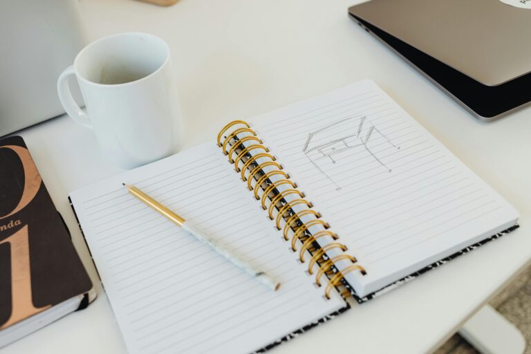 A minimalist desk setup with a spiral notebook, cup, and laptop for creative work.