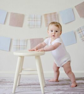 Cute baby standing by a stool indoors with a playful background.
