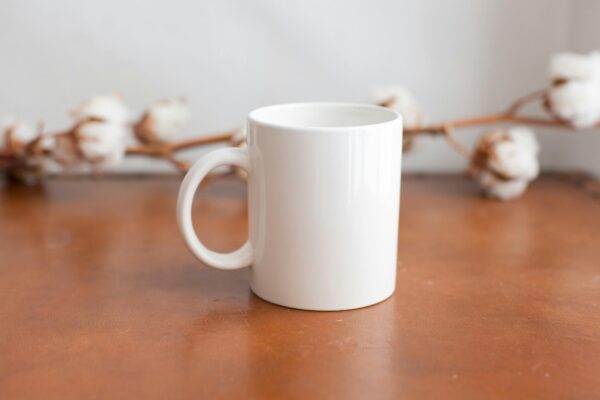 Minimalist white mug on a wooden surface with cotton flowers in the background, ideal for mockups.